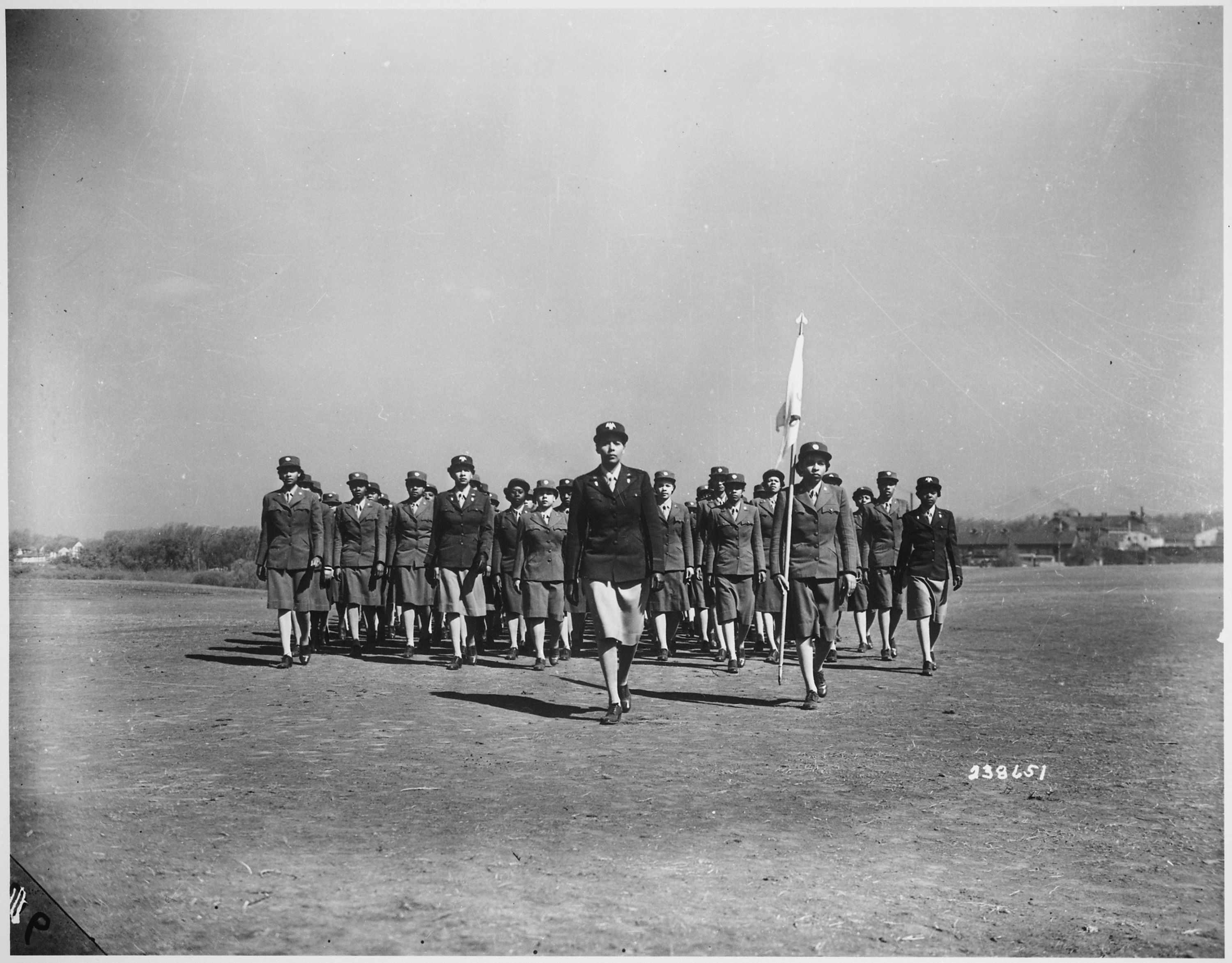 Group of female Soldiers in formation walking in a field toward the camera. One woman is in the lead with another woman to her left carrying a flag. 