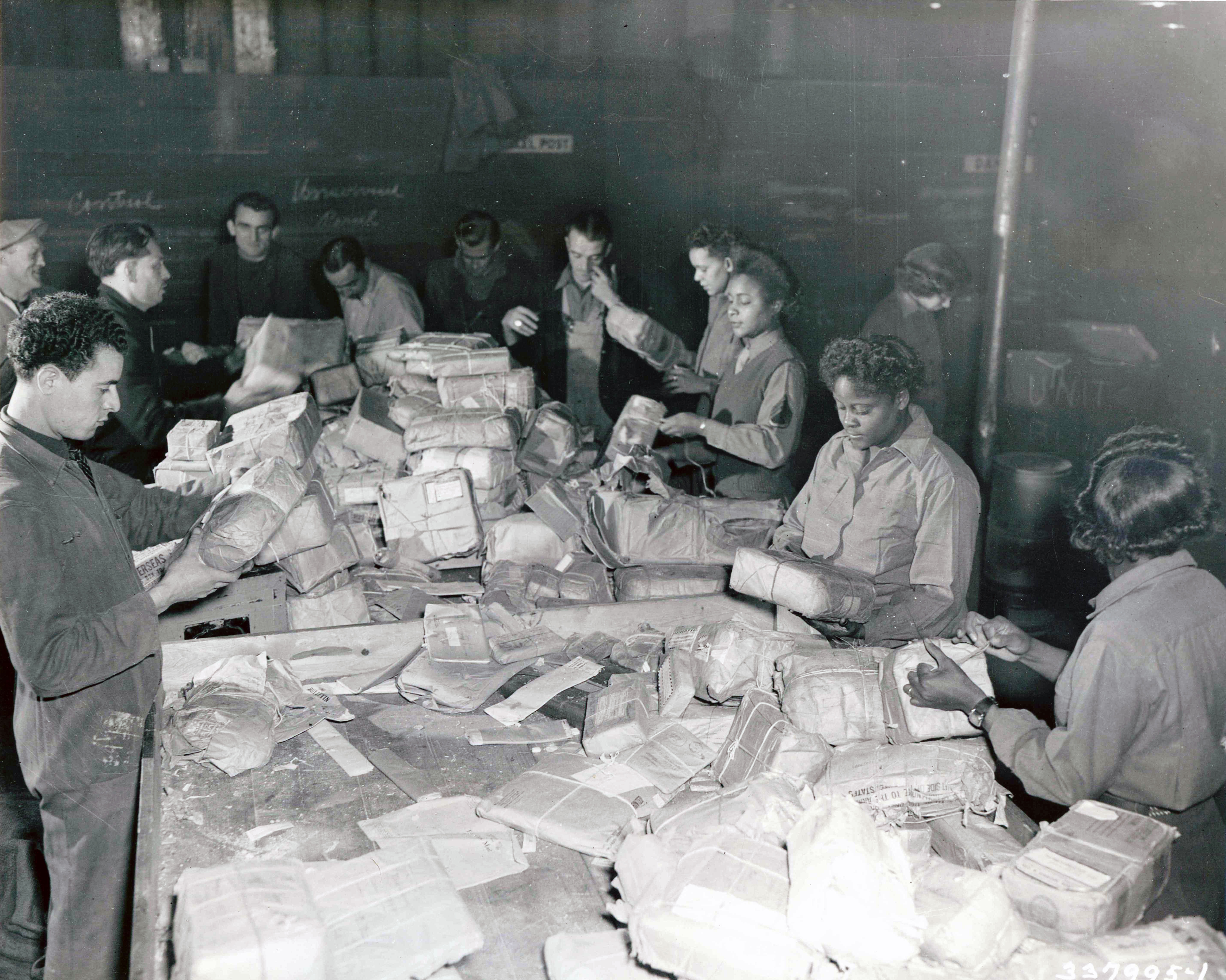 Women and men surround a large table covered in mail and packages. The are inspecting the packages in front of them. 