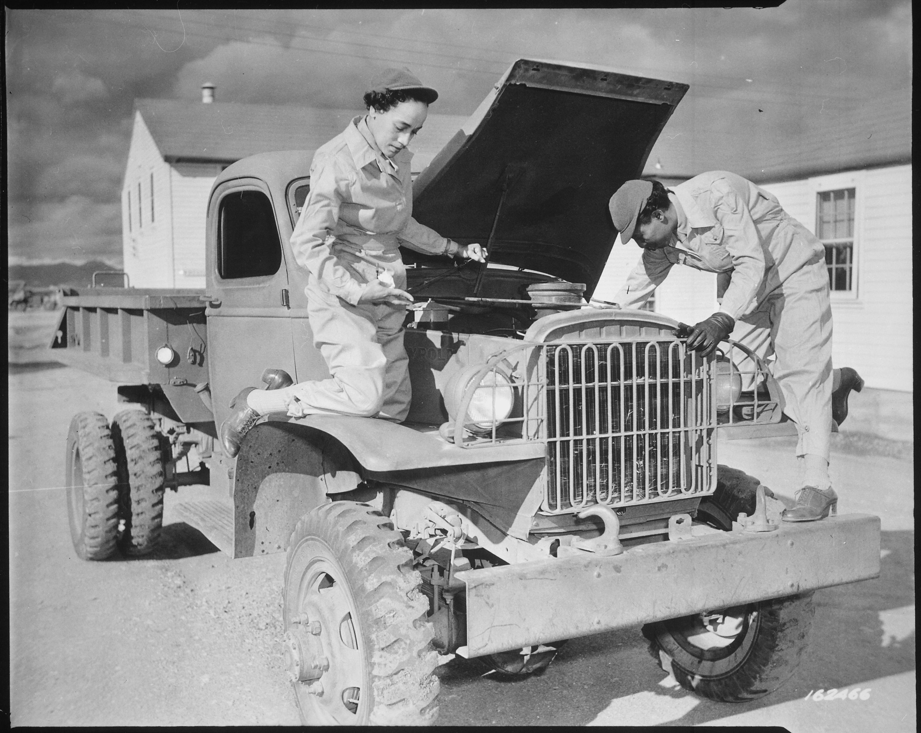 Two Black women work on a U.S. Army truck in front of a white building. 