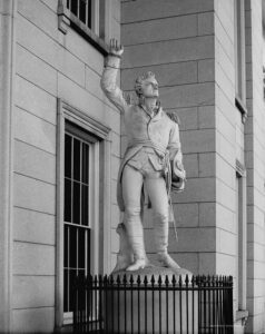 Marble statue of a man in uniform raising his right arm. A sword is hanging on his left side and he is holding his hat in his left hand. 