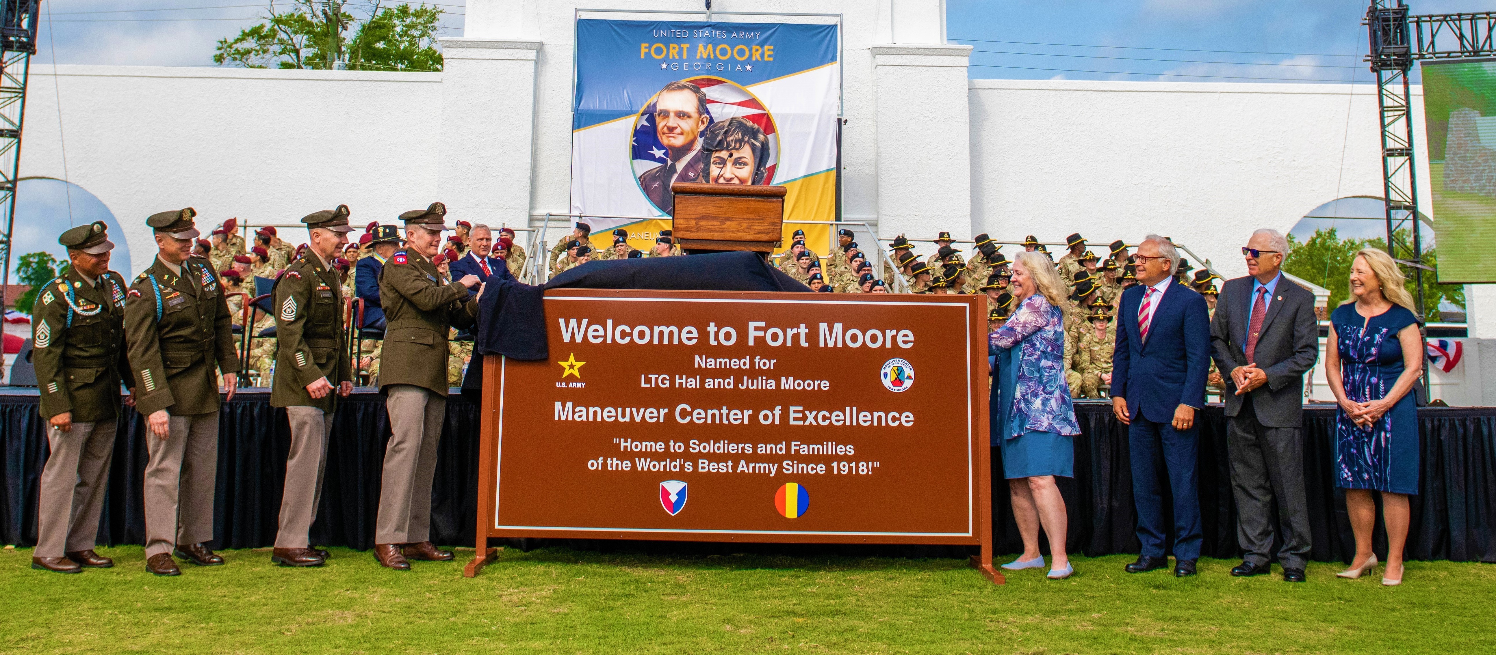 Group of Soldiers on the left and civilians on the right remove a cloth from a new sign reading "Welcome to Fort Moore. Named for LTG Hal and Julia Moore. Maneuver Center of Excellence. 'Home to the Soldiers and Families of the World's Best Army Since 1918!'"