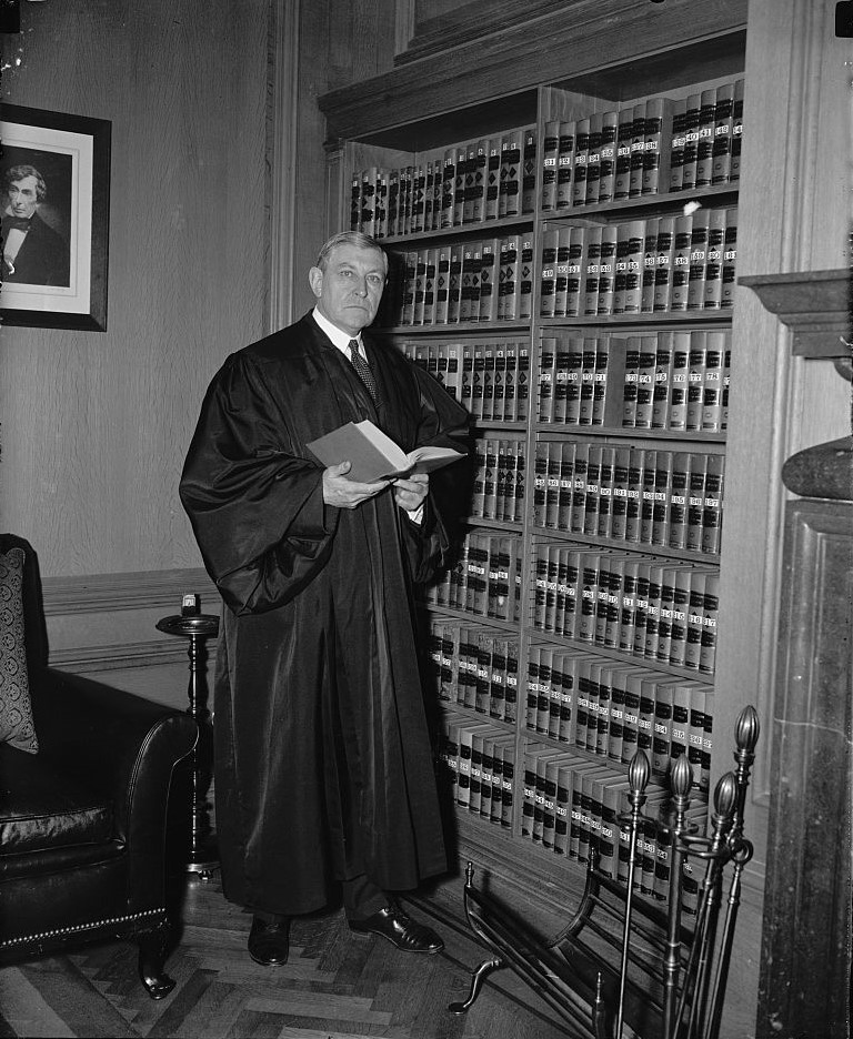 Elderly man in justice robes stands next to a book shelf holding an open book and looking at the camera. 