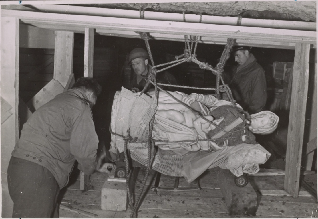 Three men examine the sculpture which is tied up with ropes and resting on a dolly