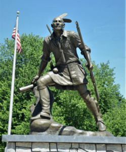 Statue of a Native American man posed with one foot on a rock and weapons in both hands.