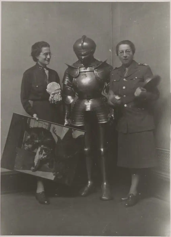 Standen is holding a sculptural head and a painting of a cat. The two women are standing on either side of a suit of armor.